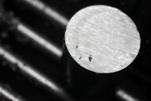 Close-up of a brushed silver metal cylinder end on a dark background, with fine woodgrain-like grooves and a few small specks on the face.
